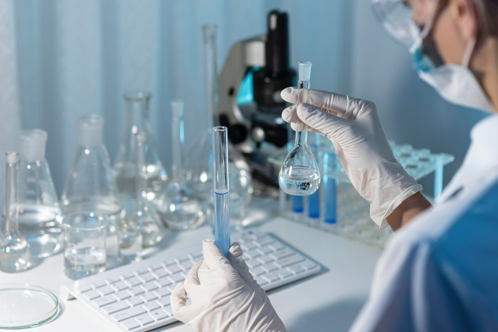 Pracownik laboratorium w rękawiczkach trzymający kolbę i probówkę z cieczą przy stanowisku badawczym